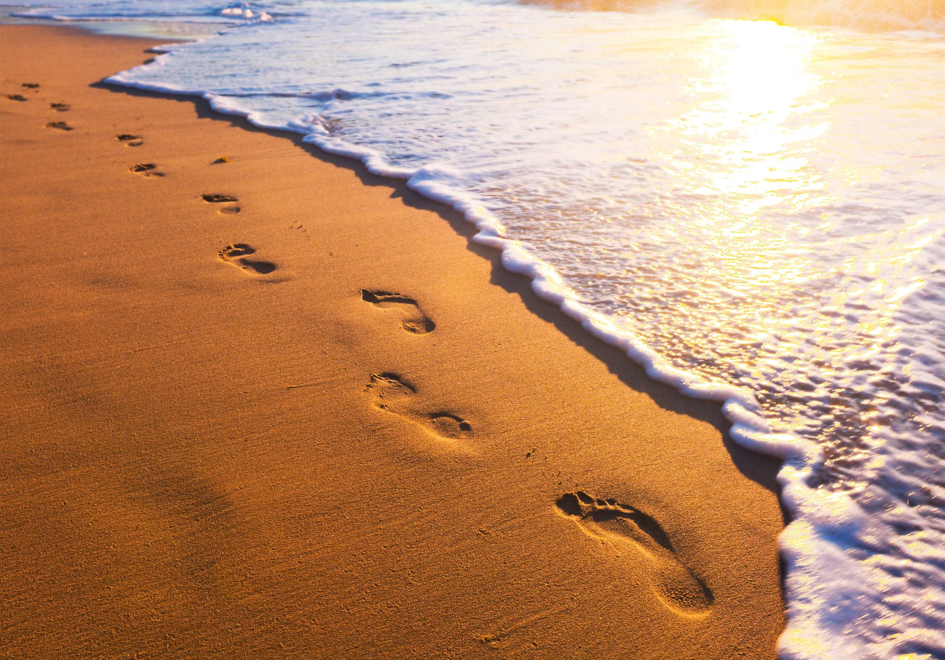 Fußabdrücke im nassen Sand am Meer, während das Sonnenlicht vom Wasser reflektiert wird.